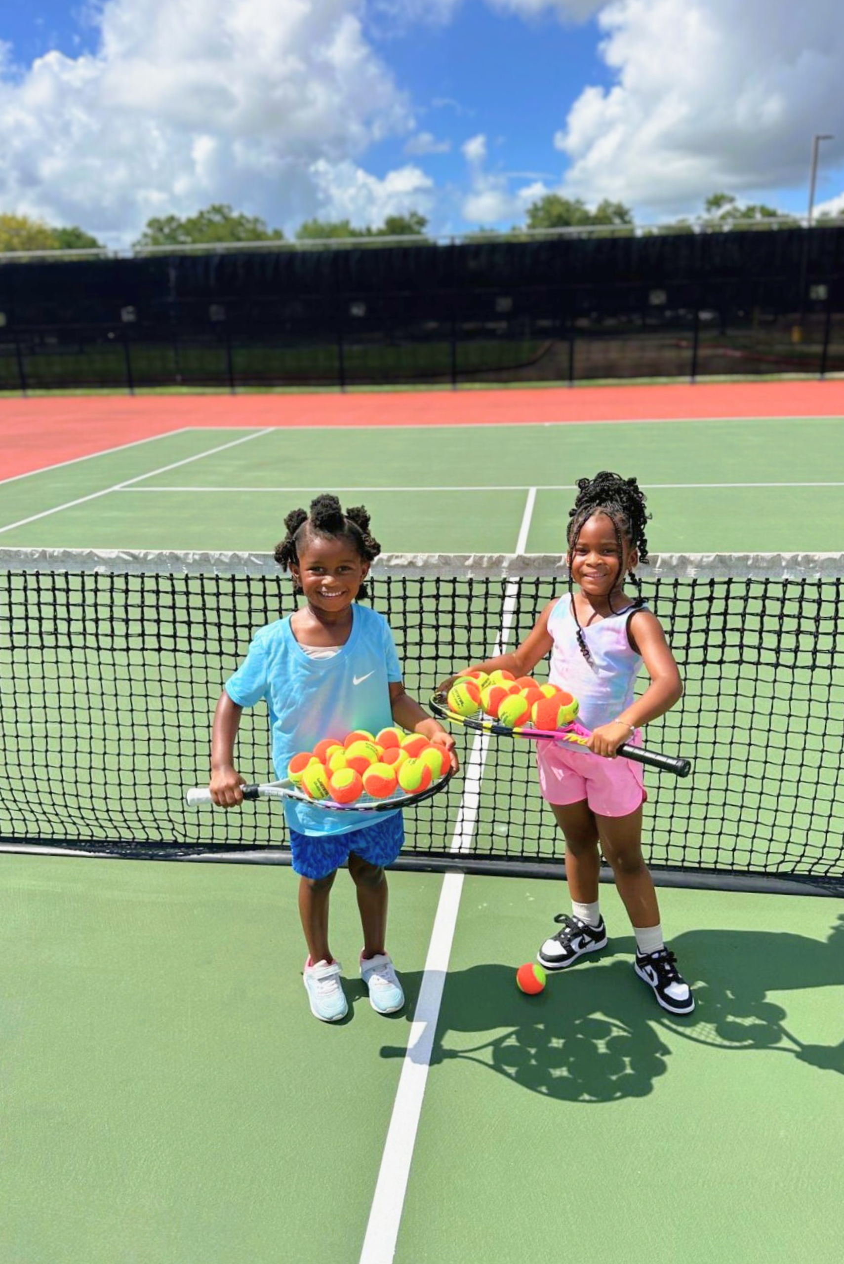 Two young girls smile happily on a tennis court while balancing dozens of tennis balls on their rackets during a fun lesson.