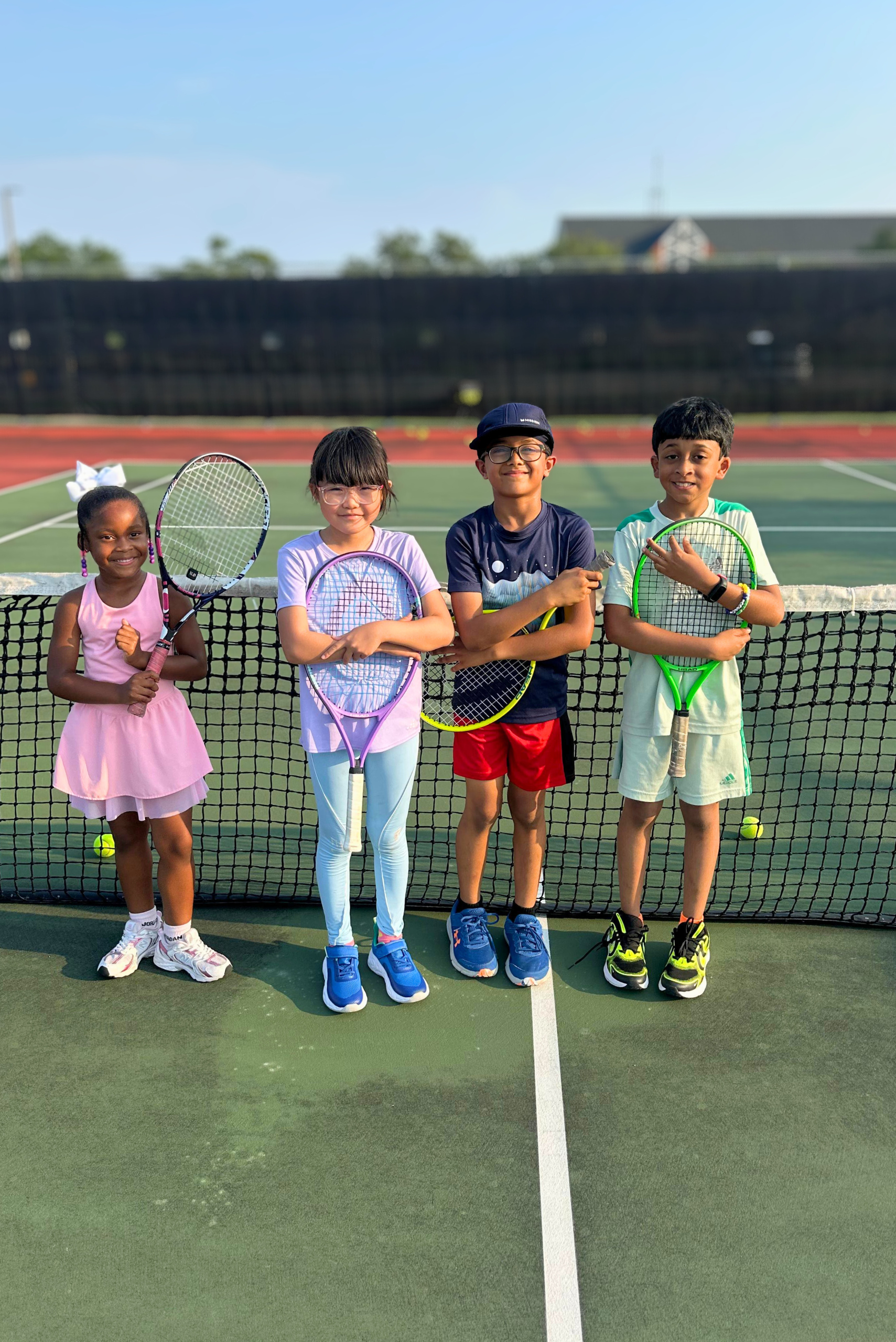 Four young, diverse children smiling proudly with their tennis rackets in front of a tennis net on a sunny court.
