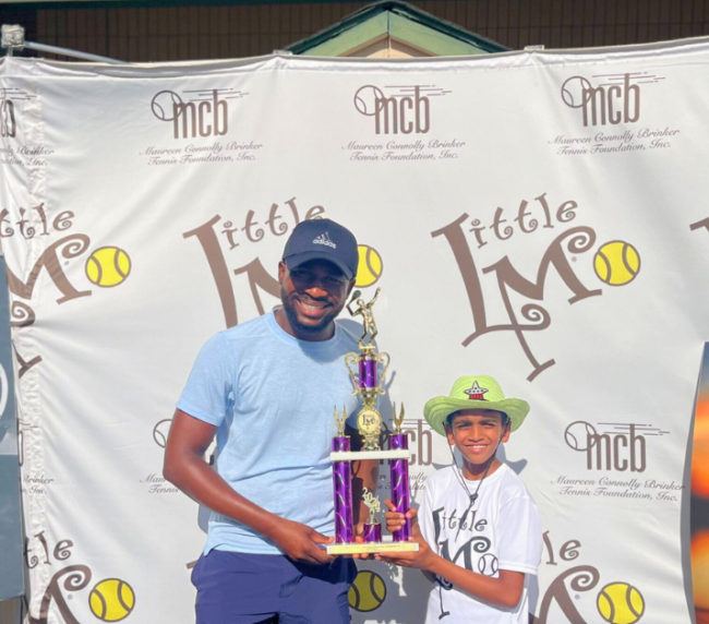 A young boy smiles proudly, holding his trophy and a sign that reads 'I'm going to USTA L1 Nationals!', in front of a table filled with trophies.