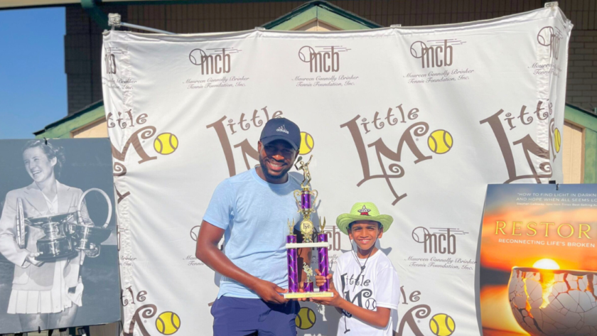 A young boy smiles proudly, holding his trophy and a sign that reads 'I'm going to USTA L1 Nationals!', in front of a table filled with trophies.