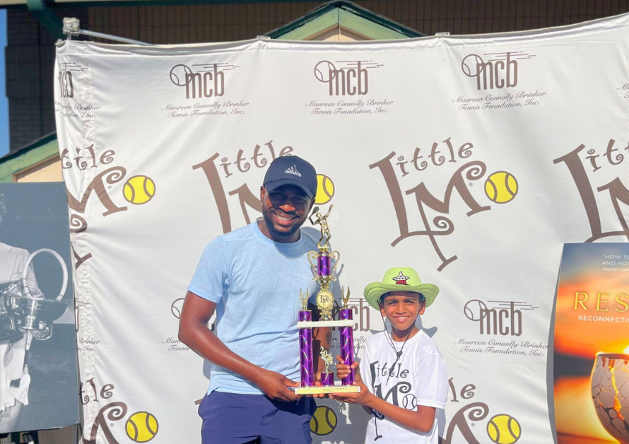 A young boy smiles proudly, holding his trophy and a sign that reads 'I'm going to USTA L1 Nationals!', in front of a table filled with trophies.