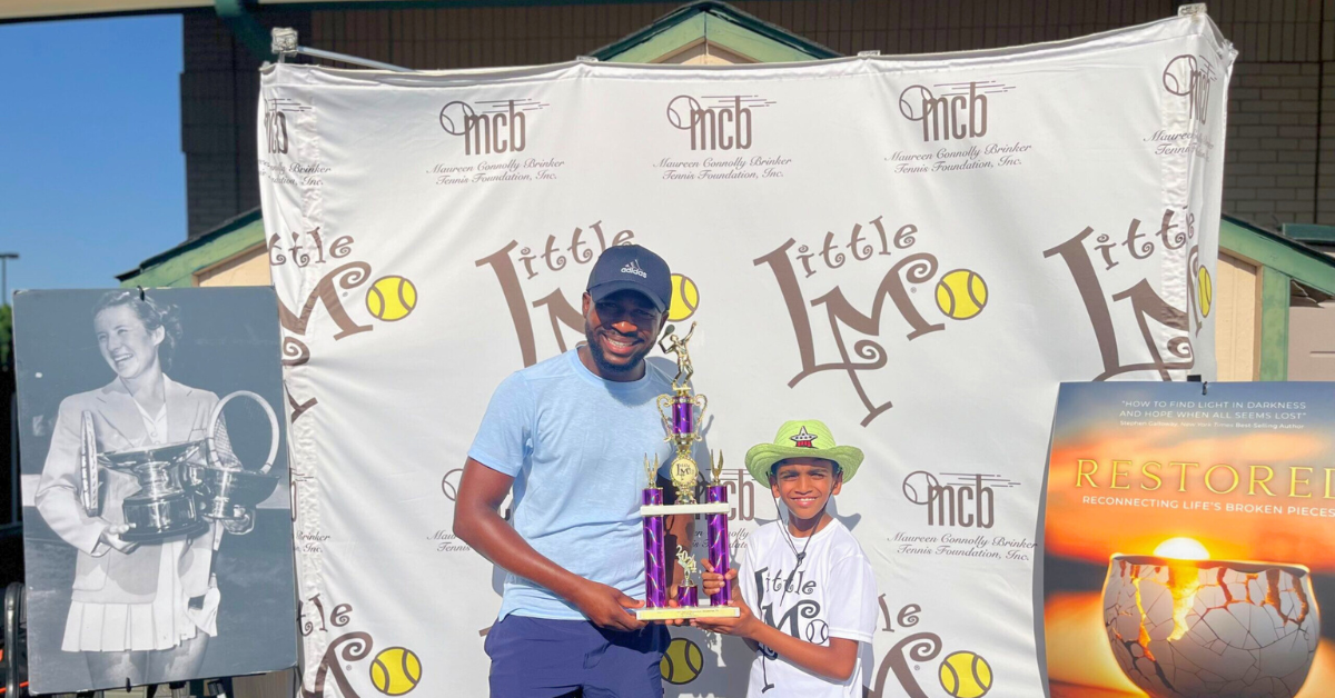 A young boy smiles proudly, holding his trophy and a sign that reads 'I'm going to USTA L1 Nationals!', in front of a table filled with trophies.